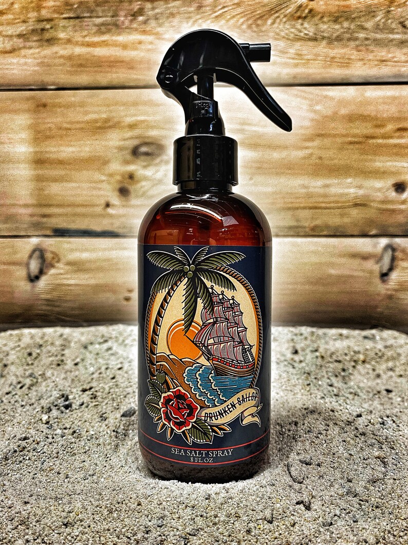 May include: A brown glass bottle with a black spray nozzle and a blue label. The label features a nautical illustration of a ship sailing on the ocean with a palm tree and a red rose. The text on the label reads "Drunken Sailor Sea Salt Spray 8 FL OZ".