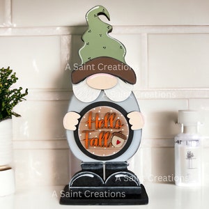 May include: A wooden gnome decoration with a green hat, brown brim, and a gray body. The gnome holds a sign that says "Hello Fall" with an acorn graphic. The base and shoes are black.