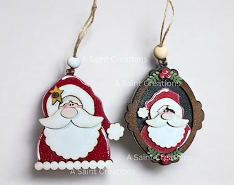 May include: Two wooden Christmas ornaments featuring Santa Claus. One ornament is a red and white Santa head with a white beard and a gold star on his hat. The other ornament is a framed Santa head with a black background and a brown frame. Both ornaments have a rustic, handmade look.