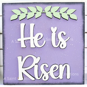 May include: A purple wooden sign with white lettering that reads "He is Risen". The sign is decorated with a green leafy wreath at the top.