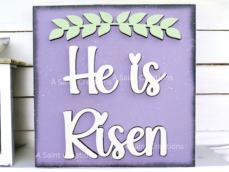 May include: A purple wooden sign with white lettering that reads "He is Risen". The sign is decorated with a green leafy wreath at the top.