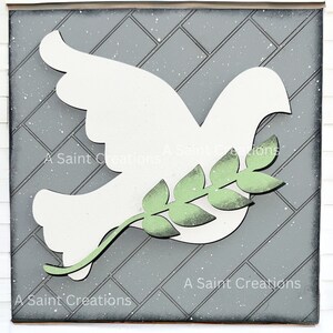 May include: A white wooden dove with an olive branch in its beak, on a gray background with a diamond pattern. The dove is a symbol of peace.