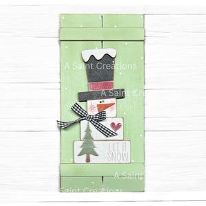 May include: A green wooden snowman decoration with a black top hat, a carrot nose, and a black and white bow. The snowman is made of stacked blocks with a heart and a Christmas tree. The text "LET IT SNOW" is visible.