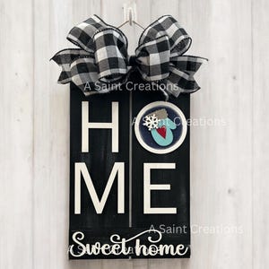 May include: Black wooden sign with the word "HOME" in white letters and the phrase "Sweet Home" in white cursive letters. A black and white checkered bow is attached to the top of the sign. A blue mitten with a red heart and snowflake is on the sign.