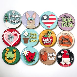 May include: Twelve round wooden signs with various seasonal and holiday themes. The signs feature painted designs and text, including "freezin season", "Feb 14", "Fresh Picked Daisies", "Back to School", "Give Thanks", "Trick or Treat", "Hello Fall", and "Santa Claus".