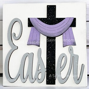 May include: A white wooden sign with the word "Easter" in gray letters and a black cross with a purple scarf draped over it. The cross and letters have a white glitter effect.