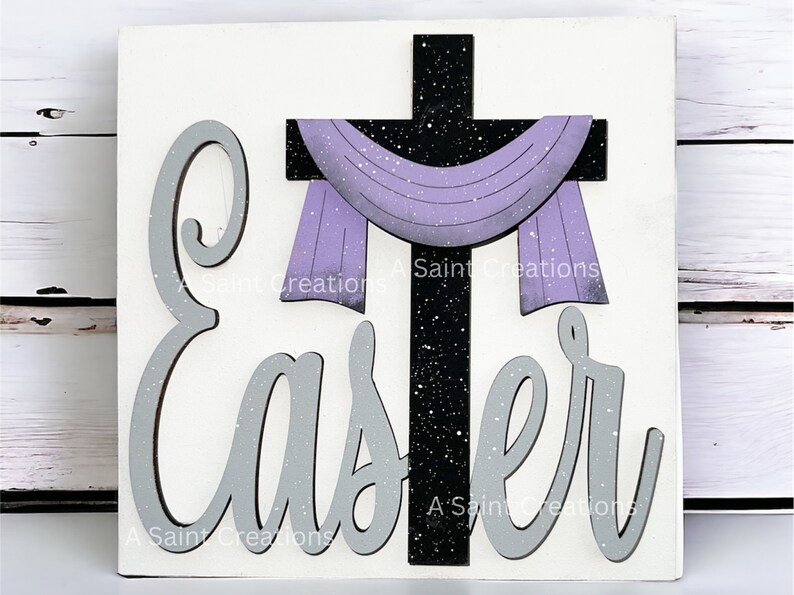 May include: A white wooden sign with the word "Easter" in gray letters and a black cross with a purple scarf draped over it. The cross and letters have a white glitter effect.