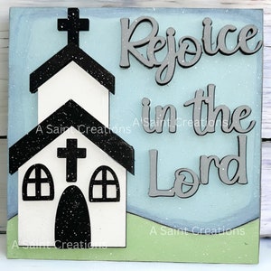 May include: A wooden sign with a blue background and a white church silhouette. The sign says "Rejoice in the Lord" in gray letters.