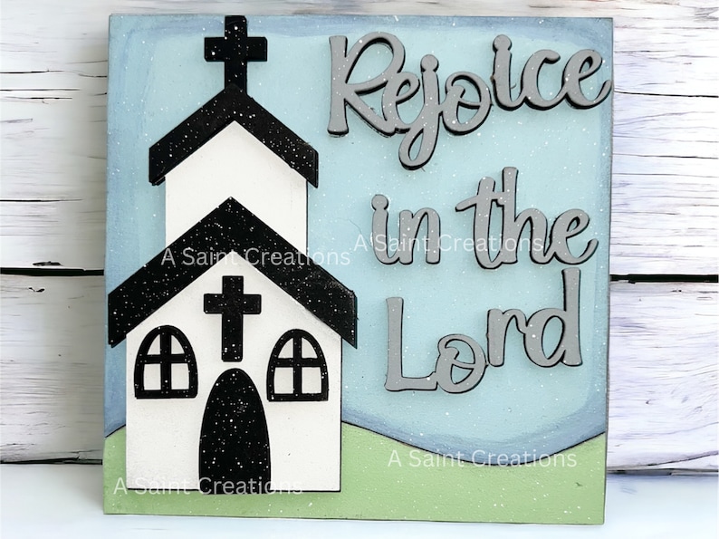 May include: A wooden sign with a blue background and a white church silhouette. The sign says "Rejoice in the Lord" in gray letters.