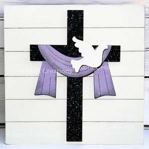 May include: A wooden cross with a black background and white speckles. A white dove is perched on the cross. A purple fabric hangs from the cross. The background is a white wood plank.