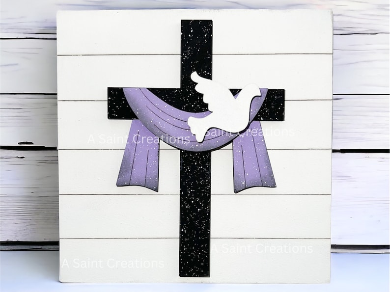 May include: A wooden cross with a black background and white speckles. A white dove is perched on the cross. A purple fabric hangs from the cross. The background is a white wood plank.