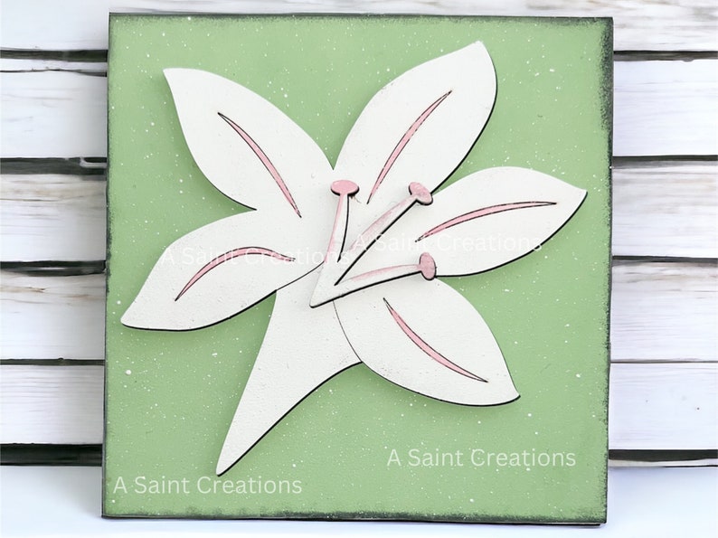 May include: A white wooden lily flower with pink accents on a green background. The flower is three-dimensional and has a layered look. The background has a speckled texture. The text "A Saint Creations" is printed at the bottom of the image.