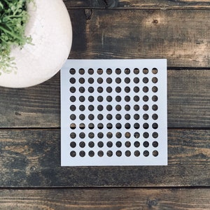 May include: White stencil with a grid of evenly spaced circles. The stencil is on a dark brown wooden surface.