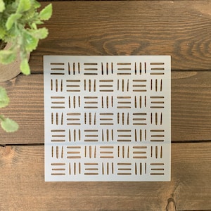 May include: A white plastic stencil with a geometric pattern of lines and dashes. The stencil is on a wooden surface.
