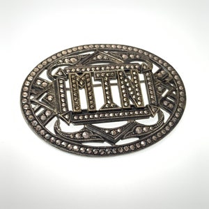 May include: A silver oval brooch with a geometric design and the letters "MTN" set in the center. The brooch is covered in small, sparkling stones.