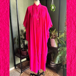 May include: A bright fuchsia, floor-length dress with a mandarin collar and button-down front. The dress has wide sleeves and is displayed on a mannequin. The dress is made of a soft, velvety fabric.