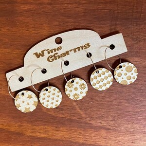 May include: Set of five wooden wine glass charms with different patterns, including stars, hearts, flowers, polka dots, and geometric shapes. The charms are attached to silver hoops and are displayed on a wooden card that reads "Wine Charms".