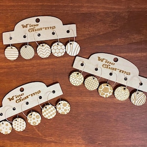 May include: Wooden wine charms with various etched patterns, including stripes, zigzags, and dots. Each charm is attached to a silver-colored ring and displayed on a wooden card that reads "Wine Charms".