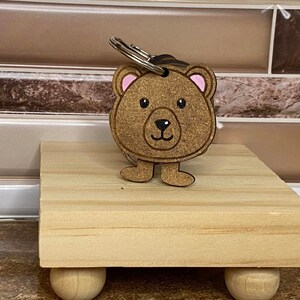 Op de afbeelding: Een houten beren sleutelhanger met roze oren en een bruin geschilderd gezicht. De sleutelhanger is bevestigd aan een metalen ring en staat op een houten standaard.