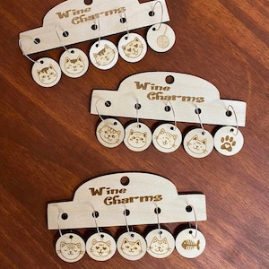May include: Set of wooden wine glass charms with cat and paw print designs. The charms are attached to silver rings and are displayed on a wooden board with the text "Wine Charms".