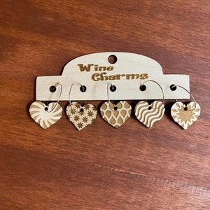 May include: A set of five wooden heart-shaped wine charms with various etched designs, including stripes, stars, and a sunburst. The charms hang from silver hoops and are attached to a wooden display card that reads "Wine Charms".