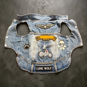 May include: A light blue acid-wash denim vest for pets featuring a collar, armholes, and snap closures. Patches include a winged skull, a motorcycle, a paw print, a skull and crossbones, a yellow brand logo, and a black patch reading LONE WOLF.