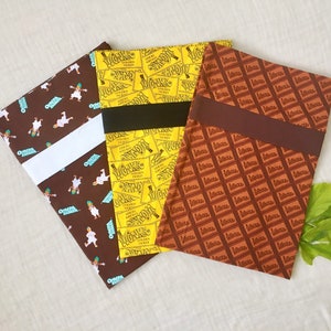 May include: Three fabric book covers with different patterns. The first cover is brown with a white stripe and a repeating pattern of a person in a blue and white outfit. The second cover is yellow with a repeating pattern of a chocolate bar with the text "Wonka Golden Ticket". The third cover is brown with a repeating pattern of a chocolate bar with the text "Wonka".