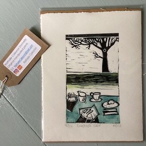 May include: A black and white linocut print of a roadside cafe with a tree in the background. The print is titled "Roadside Cafe" and is numbered 16/30.