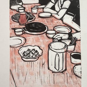 May include: A black and white linocut print of a table setting with a checkered tablecloth. The table is set with plates, cups, and a teapot. There is a bowl of food in the center of the table.