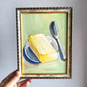 May include: Oil painting of a yellow butter block on a blue plate with a silver butter knife. The painting is in a gold-colored ornate frame. The background is light green.