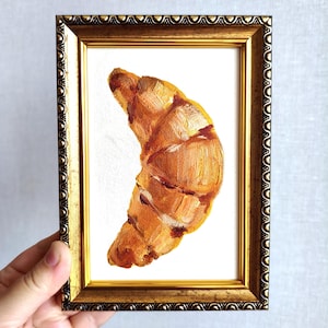 May include: An oil painting of a golden-brown croissant on a white background, encased in a rectangular, ornate, metallic gold frame with a textured, scalloped edge. The brushstrokes are visible, highlighting the flaky layers of the pastry.