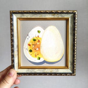 May include: A framed painting of a halved hard-boiled egg with a bright yellow yolk and sprinkled seasonings. The egg is set against a light gray background, within a gold and white ornate frame.