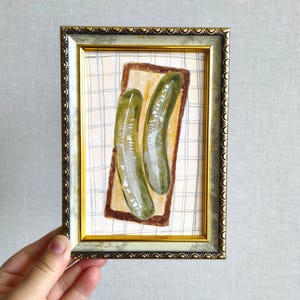 May include: A framed watercolor painting featuring two green pickles resting on a slice of brown bread. The background consists of a light-colored fabric with a subtle blue and white grid pattern. The frame is ornate with gold and silver tones.