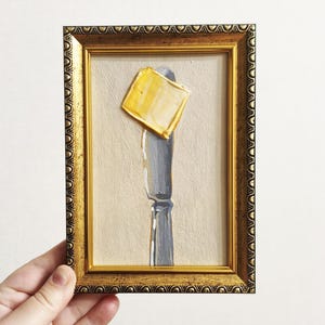 Può includere: Dipinto incorniciato di un coltello da burro con un quadrato di burro. Il coltello è grigio e il burro è giallo. La cornice è dorata con dettagli ornati. Lo sfondo è beige.
