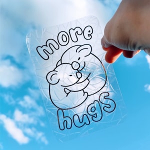 Puede incluir: Una pegatina transparente con un efecto de vidrio agrietado que presenta dos koalas abrazándose. La pegatina dice "more hugs" en texto negro. La pegatina mide 10x15 cm.