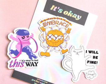 May include: A collection of stickers with whimsical designs and text. One sticker features a running figure with the text "It's okay to feel this way." Another shows a flower with the words "Embrace." A cat sticker says "I will be fine!" with "Doubt" below.
