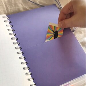 May include: A spiral-bound notebook with a purple cover and pocket pages. The notebook is open, revealing white grid paper and a purple pocket holding a colorful card. The text "Pocket pages" is at the top.