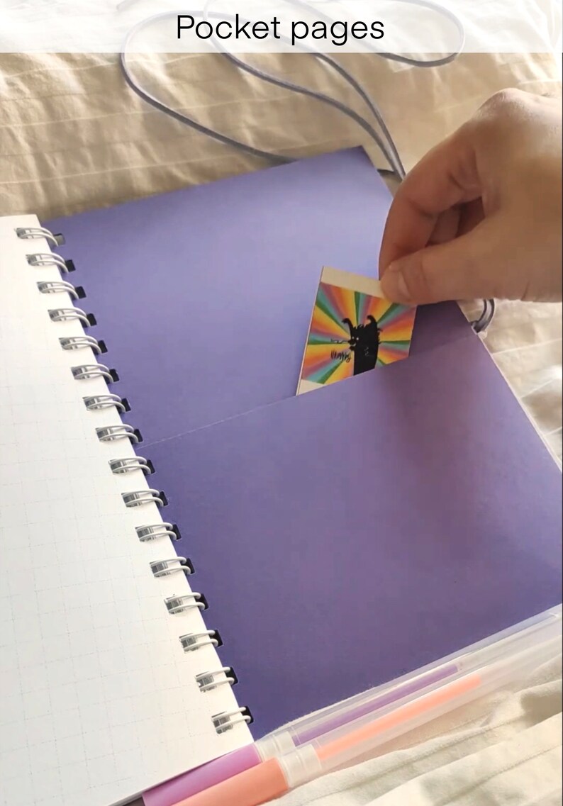 May include: A spiral-bound notebook with a purple cover and pocket pages. The notebook is open, revealing white grid paper and a purple pocket holding a colorful card. The text "Pocket pages" is at the top.