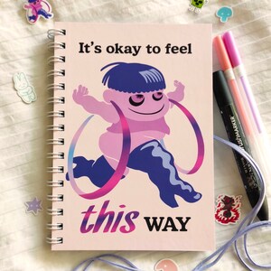 May include: Spiral-bound notebook with a pink cover featuring a cartoon figure and the text "It's okay to feel this WAY." The notebook has 170 pages of 80gsm high-quality stock. Pens and stickers are also visible.