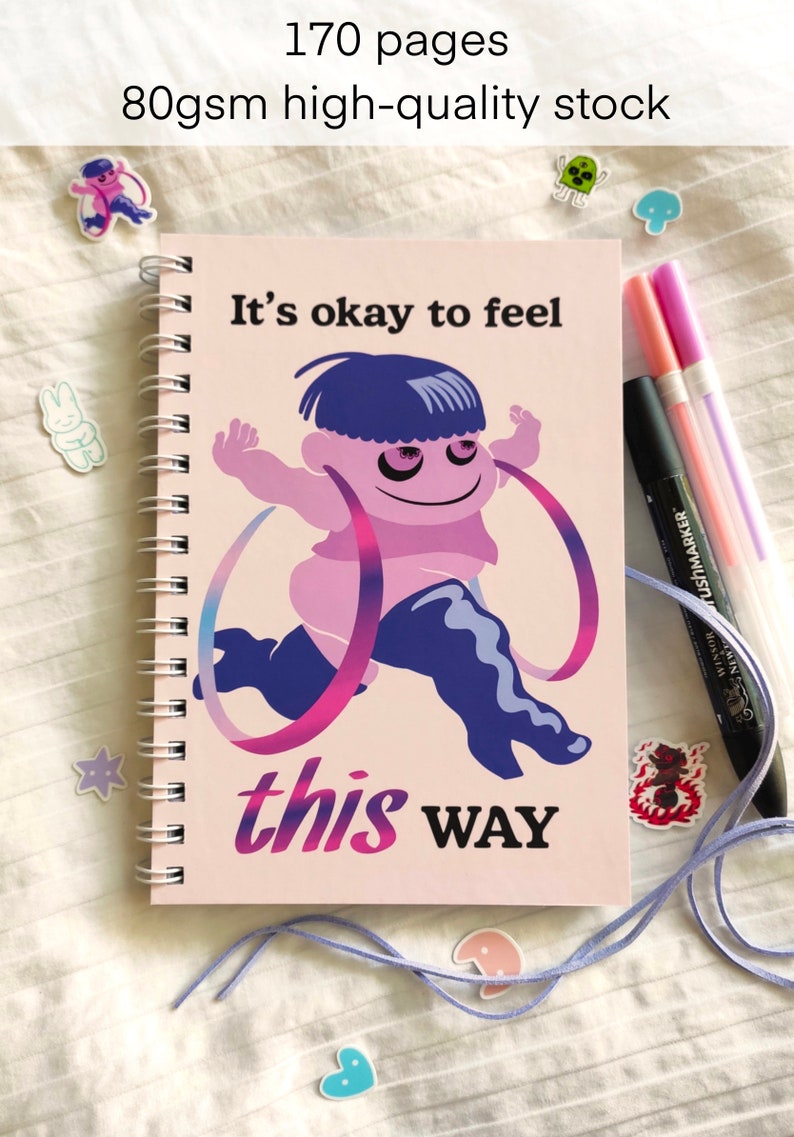 May include: Spiral-bound notebook with a pink cover featuring a cartoon figure and the text "It's okay to feel this WAY." The notebook has 170 pages of 80gsm high-quality stock. Pens and stickers are also visible.