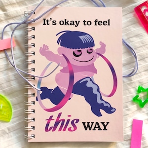 May include: Spiral-bound notebook with a cartoon illustration of a running figure with the text "It's okay to feel this WAY." The notebook is A5 size, approximately 5-7/8 x 8-1/4 inches. The cover art features purple and pink hues.