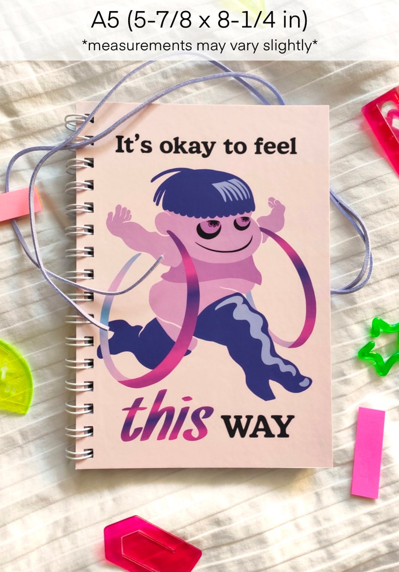 May include: Spiral-bound notebook with a cartoon illustration of a running figure with the text "It's okay to feel this WAY." The notebook is A5 size, approximately 5-7/8 x 8-1/4 inches. The cover art features purple and pink hues.