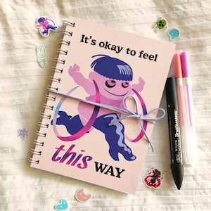 May include: A spiral-bound notebook with the text "It's okay to feel this way" and an illustration of a running figure. The notebook is tied with a lavender ribbon. Also shown are several stickers and three pens.