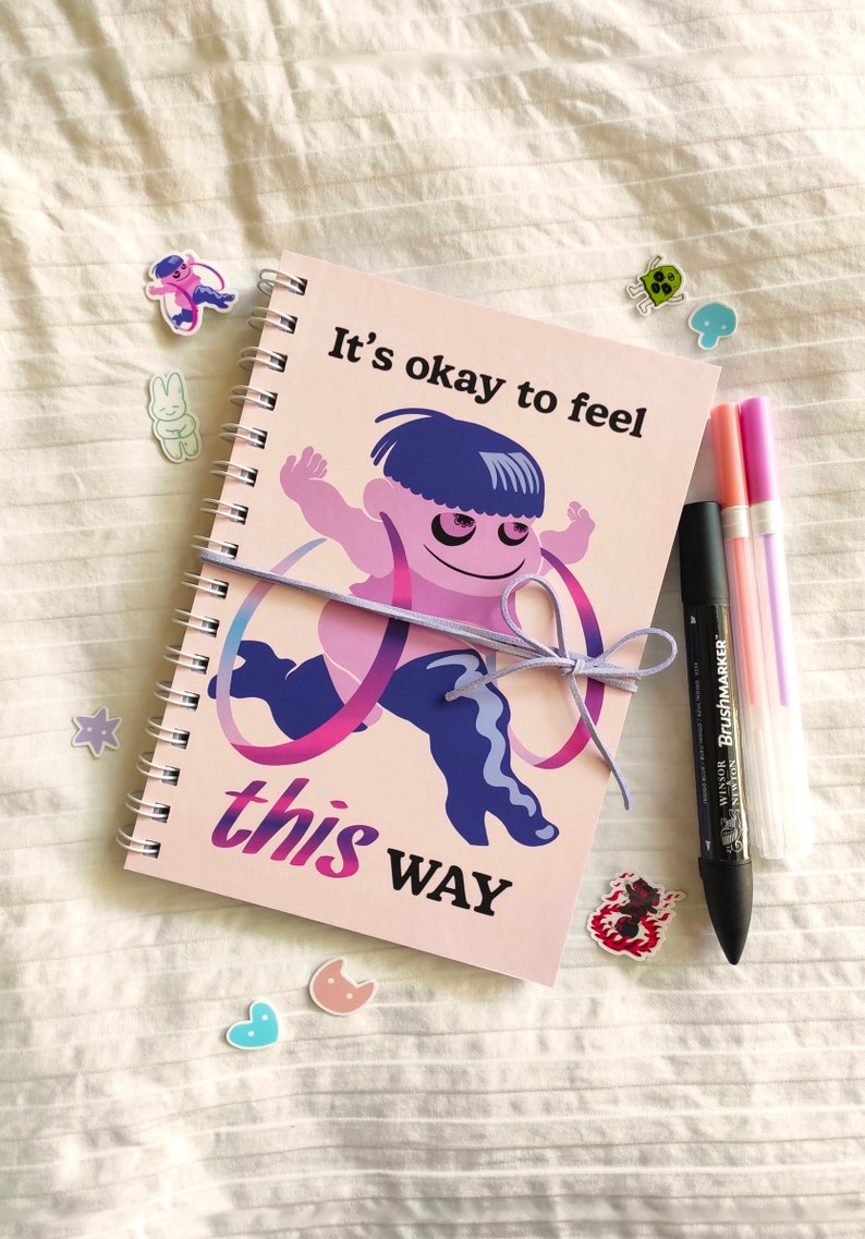 May include: A spiral-bound notebook with the text "It's okay to feel this way" and an illustration of a running figure. The notebook is tied with a lavender ribbon. Also shown are several stickers and three pens.