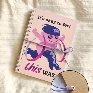 May include: A spiral-bound notebook with a pink cover featuring a cartoon figure and the text "It's okay to feel this WAY." The notebook is closed with a faux leather string closure.