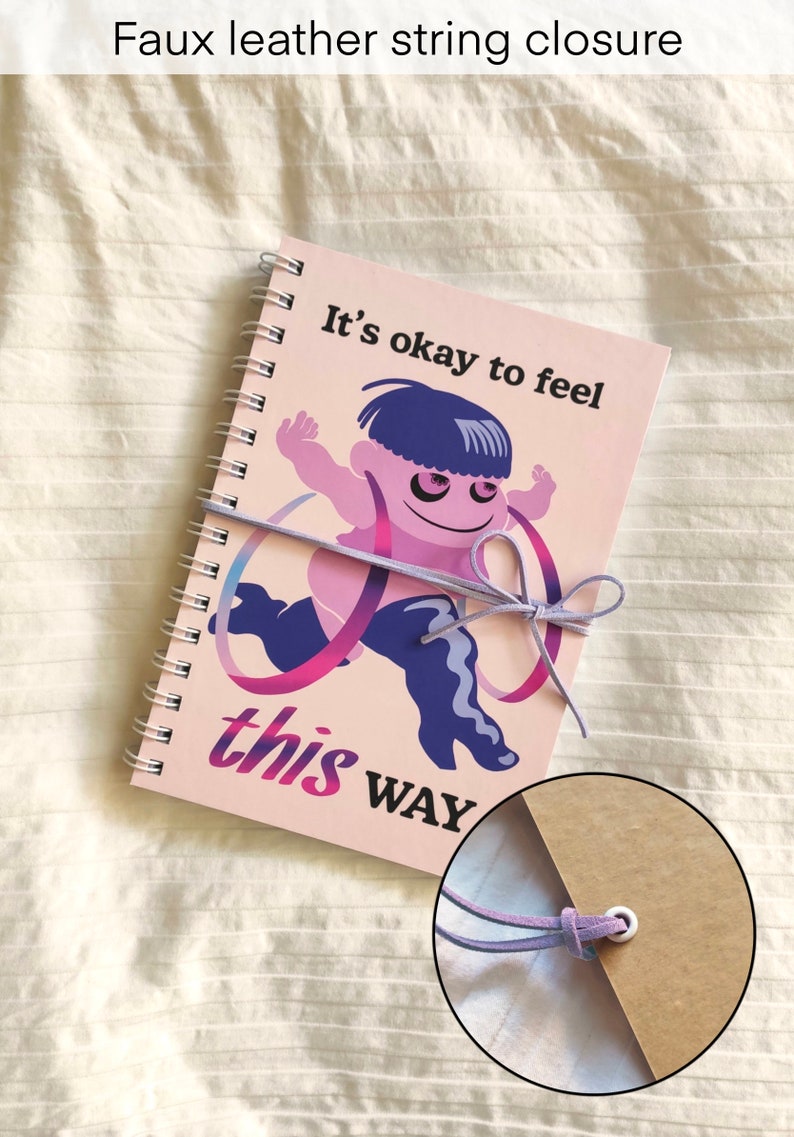May include: A spiral-bound notebook with a pink cover featuring a cartoon figure and the text "It's okay to feel this WAY." The notebook is closed with a faux leather string closure.