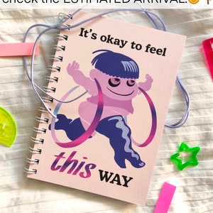 May include: A spiral-bound notebook with a pink cover featuring a cartoon figure and the text "It's okay to feel this way." The figure is purple and pink, with blue hair and boots. A light purple cord is attached.