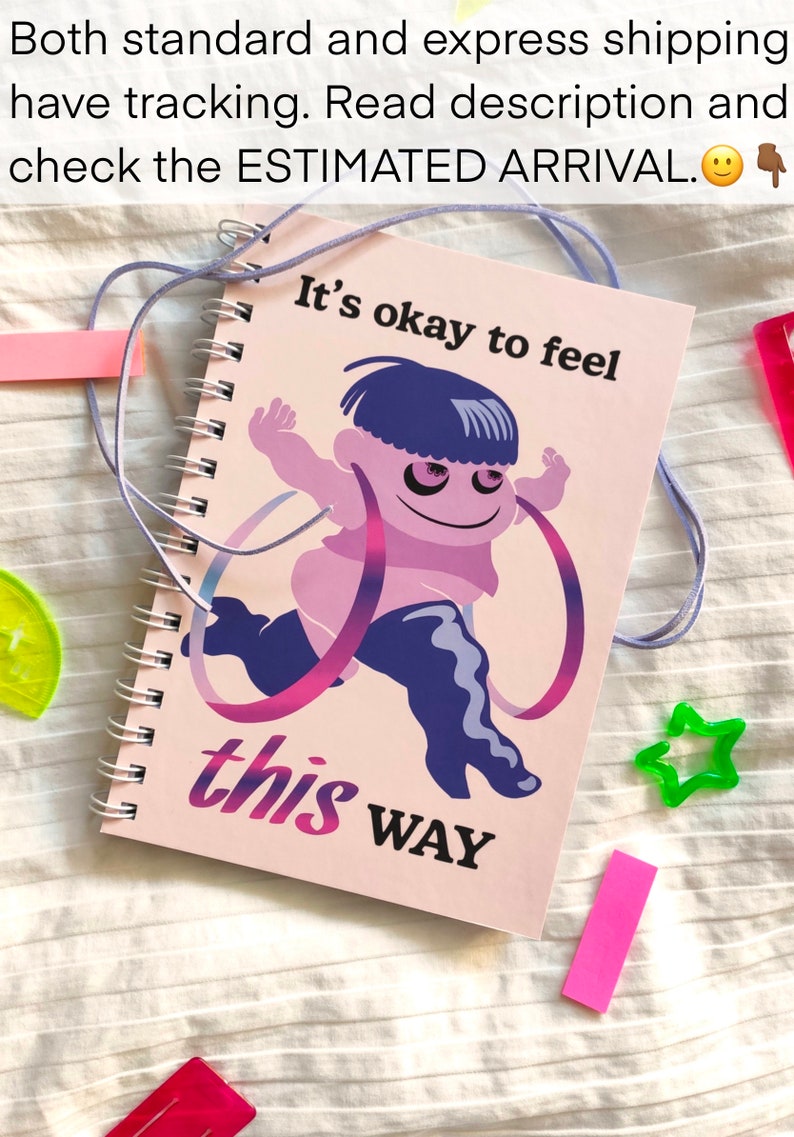 May include: A spiral-bound notebook with a pink cover featuring a cartoon figure and the text "It's okay to feel this way." The figure is purple and pink, with blue hair and boots. A light purple cord is attached.