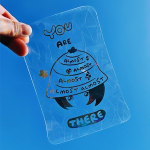 May include: A clear, rectangular sticker with the words "YOU ARE" above a cartoon design and the word "THERE" below. The cartoon design includes the word "ALMOST" multiple times. The sticker measures 3.93 x 5.9 inches.