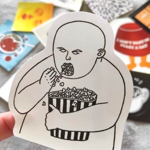 May include: A white sticker featuring a black and white illustration of a person eating popcorn from a striped container. The sticker has the text "Journal stickers included in each order" at the top.